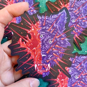 Op de afbeelding: Een patch met een gesplitst ontwerp van een wolfsgezicht. Eén kant is paars, de andere is rood met een burst-effect. De wolf heeft scherpe tanden en is omgeven door rode spikes. De patch heeft een zwarte achtergrond.