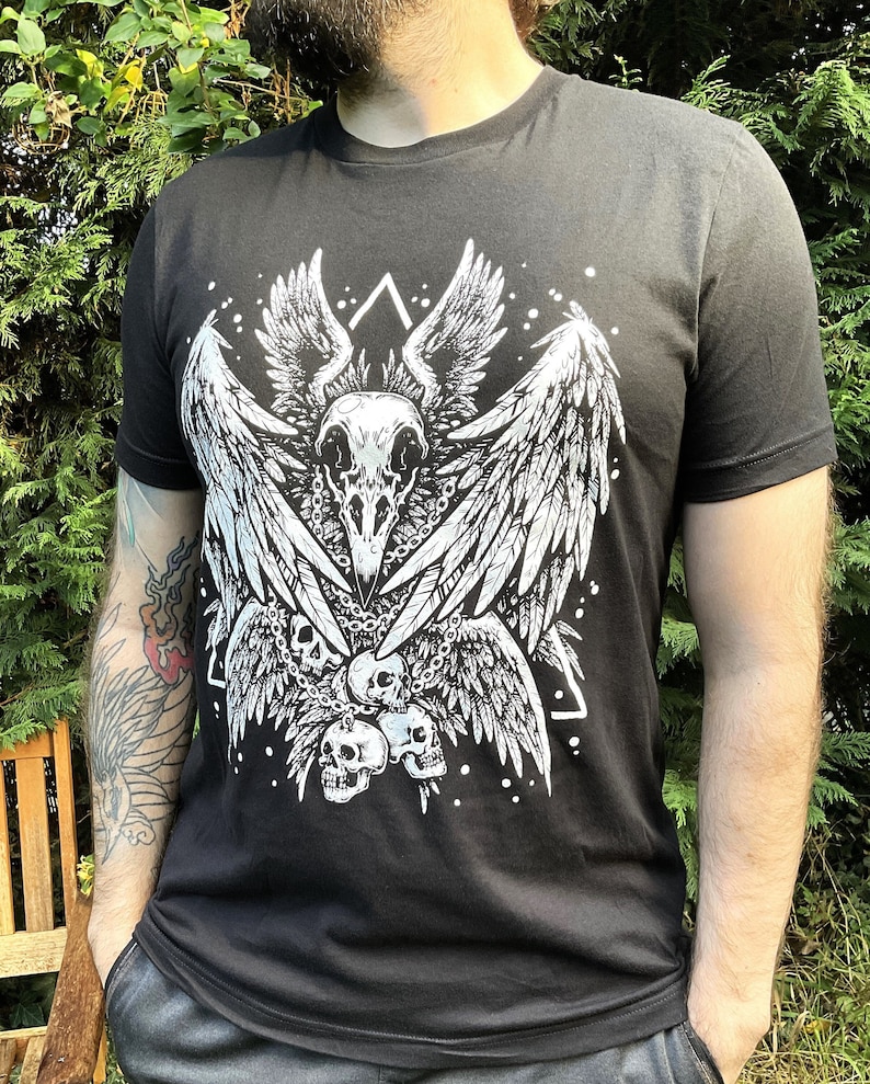 Peut inclure: T-shirt noir avec un imprim&eacute; blanc d'un cr&acirc;ne avec des ailes et des cha&icirc;nes. Le cr&acirc;ne est entour&eacute; d'un triangle et de petits points blancs.