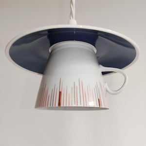 May include: A unique pendant light fixture made from a white teacup and saucer. The saucer is navy blue with a white rim, and the cup features vertical lines in shades of brown and gold. The light hangs from a white cord.