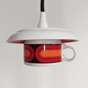 May include: A unique pendant light fixture made from a vintage coffee mug. The mug features a retro design with red, orange, and black geometric shapes. The light has a white shade and a brown cord, creating a stylish and functional piece.