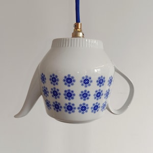 Könnte beinhalten: Eine weiße Keramik-Teekanne, die zu einer Pendelleuchte umfunktioniert wurde. Die Teekanne hat ein blaues Blumenmuster und ein blaues, stoffummanteltes Kabel. Die Lampenfassung ist goldfarben.