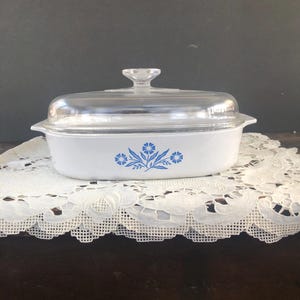 May include: White rectangular casserole dish with a clear glass lid. The dish features a blue floral design on the front. The lid has a small handle. The dish is sitting on a white lace doily.