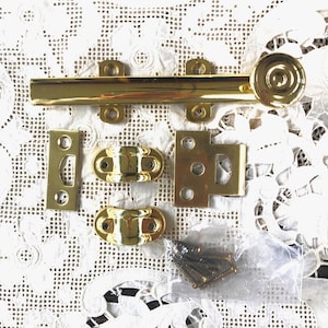 May include: A collection of shiny brass hardware components, including a long sliding bolt, various mounting plates, and screws. The pieces are arranged on a white lace surface, suggesting a vintage or antique theme.