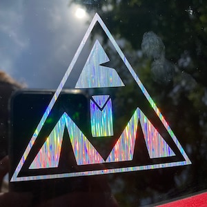 May include: A silver holographic sticker with a triangle design. The triangle contains a stylised mountain range with a single tree in the centre.