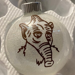 Könnte beinhalten: Ein weißer gläserner Weihnachtsbaumschmuck mit einem braunen Glitzerdesign einer Kreatur mit großen Ohren und einer langen Schnauze. Der Schmuck hat eine silberne Spitze.