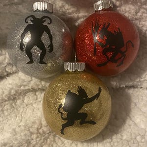 Puede incluir: Tres adornos navideños brillantes. Uno plateado, uno rojo y uno dorado. Cada adorno presenta una silueta negra de una criatura con cuernos. Los adornos tienen tapas metálicas plateadas para colgar.