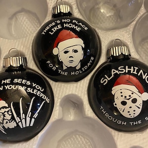 Könnte beinhalten: Vier schwarze Weihnachtskugeln mit weißem Text und roten und weißen Bildern von Figuren aus Horrorfilmen. Die Kugeln zeigen "There's no place like home for the holidays", "He sees you when you're sleeping", "Slashing through the snow" und eine Figur mit einer Maske.