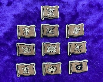 May include: A collection of ten wooden pirate flag pins with various skull and crossbones designs. Each flag is brown with a silver border and a unique skull illustration. The pins are arranged on a purple velvet surface.