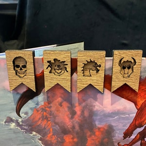 May include: Four wooden bookmarks with different designs. The bookmarks are shaped like banners and feature a skull, a monster, a dragon, and a creature with horns. The bookmarks are displayed against a background of a fantasy illustration.