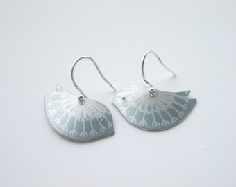 May include: A pair of silver bird-shaped earrings. Each earring features a light blue body with a white, stylized floral design. The earrings have curved silver ear wires for pierced ears.
