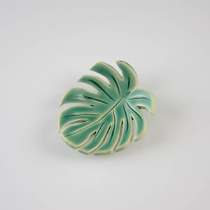 May include: A ceramic leaf dish in the shape of a monstera leaf. The leaf is green with a light green outline.