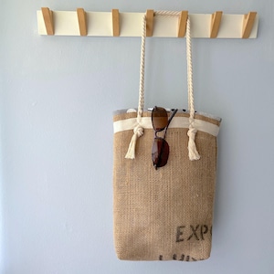 May include: A tan hessian tote bag with a cream-coloured fabric band at the top and thick white rope handles. The bag features black printed text reading EXP and is hanging from a wooden wall hook. A pair of dark brown sunglasses rests on the bag.
