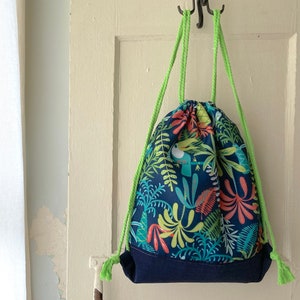 Peut inclure: Un sac à cordon bleu avec un imprimé tropical composé de feuilles vertes, de fleurs rouges et d'oiseaux bleus. Le sac a des cordons de serrage verts et un fond bleu marine.
