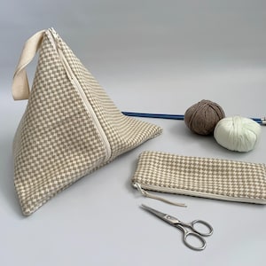 May include: A selection of craft items. A beige and white checked triangular knitting project bag, a matching pencil case, two balls of yarn, knitting needles, and scissors are visible.