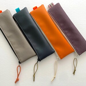 Peut inclure: Quatre étuis à crayons rectangulaires de couleurs et textures différentes. Les étuis sont gris, bleu marine, orange et violet. Chaque étui a une fermeture éclair et une petite boucle à l'extrémité. Les étuis ont une petite languette de couleur contrastante.