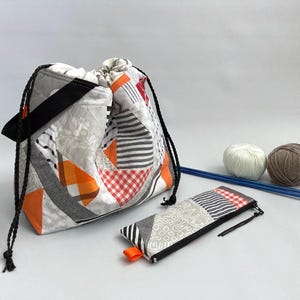 Peut inclure: Un sac à cordon en patchwork blanc et orange avec une sangle noire et une petite pochette assortie. Le sac est rempli de laine et d'aiguilles à tricoter.