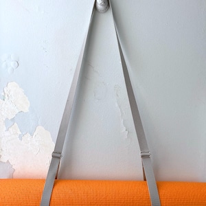 Könnte beinhalten: Ein grauer Yogamatte-Gurt, der an einem weißen, hasenförmigen Haken an einer weißen Wand hängt. Der Gurt hält eine orangefarbene Yogamatte.