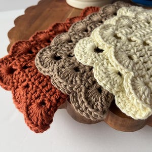 K&ouml;nnte beinhalten: Vier geh&auml;kelte Untersetzer in verschiedenen Braunt&ouml;nen, Beige und Orange. Die Untersetzer sind mit einem gewellten Rand und einem zarten Lochmuster gearbeitet.