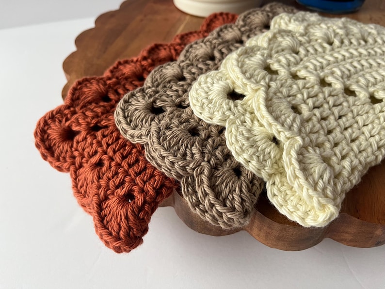 K&ouml;nnte beinhalten: Vier geh&auml;kelte Untersetzer in verschiedenen Braunt&ouml;nen, Beige und Orange. Die Untersetzer sind mit einem gewellten Rand und einem zarten Lochmuster gearbeitet.