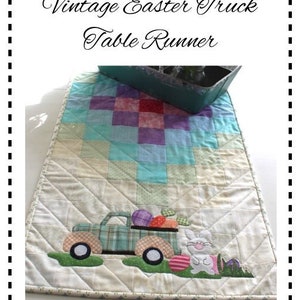 May include: A white quilted table runner with a patchwork design in shades of blue, green, purple, and pink. A vintage truck with Easter eggs is appliqued on the runner, along with a bunny rabbit. The text "Vintage Easter Truck Table Runner" is printed above the runner.