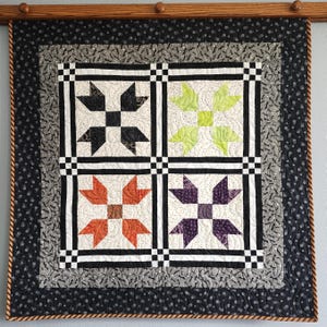 May include: A square quilt with a wooden hanging rod. The quilt features four blocks with star-like designs in black, green, orange, and purple. The blocks are set against a white background, framed by black and white checkered borders.