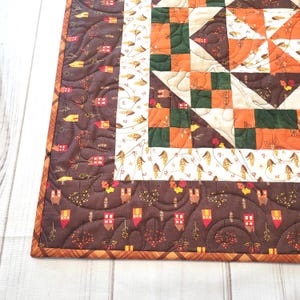 May include: A quilted fabric piece featuring a patchwork design with autumn colors including brown, orange, green, and cream. The border displays a pattern of small houses and trees, while the center contains geometric shapes and leaf motifs.