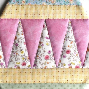 May include: A pink and white quilted fabric with a geometric pattern of triangles. The fabric has a floral print and is sewn together with a white thread.