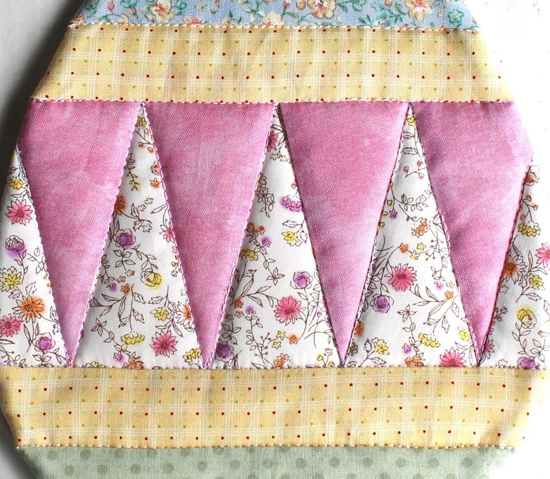 May include: A pink and white quilted fabric with a geometric pattern of triangles. The fabric has a floral print and is sewn together with a white thread.