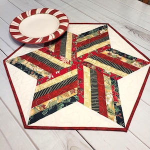 May include: A star-shaped quilted placemat with a red border and a cream-colored background. The star design features strips of red, black, gold, and floral patterned fabric. A red and white striped plate rests on the placemat, suitable for holiday dining.
