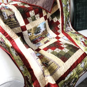 May include: A patchwork quilt featuring a farm scene with red barns, white houses, and green fields. The quilt has a cream-colored background with red and green accents. The quilt is folded over a white surface.