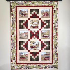 May include: A quilted wall hanging featuring a patchwork design with squares of red, green, and cream. The center showcases framed scenes of rural landscapes with red barns and covered bridges. The border has a repeating pattern of houses and trees.