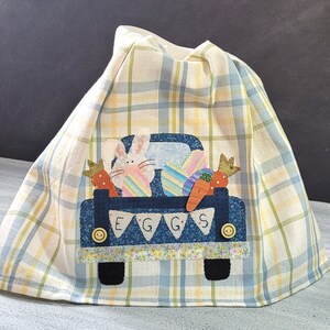 May include: A blue and white checkered kitchen towel with an embroidered design of a blue pickup truck carrying Easter eggs, a bunny, and carrots. The truck has a banner that reads "EGGS".