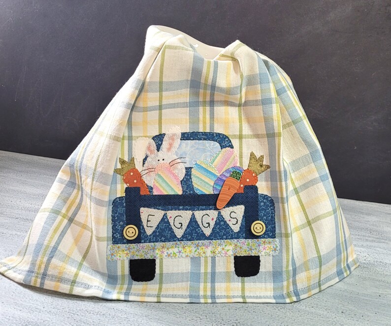 May include: A blue and white checkered kitchen towel with an embroidered design of a blue pickup truck carrying Easter eggs, a bunny, and carrots. The truck has a banner that reads "EGGS".