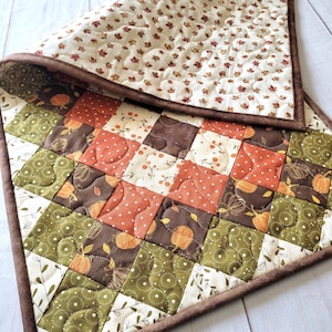 May include: A quilted table runner featuring a patchwork design of squares in shades of orange, brown, and olive green. The fabric patterns include pumpkins, berries, and leaves. The runner has a dark brown border and a cream-colored backing.