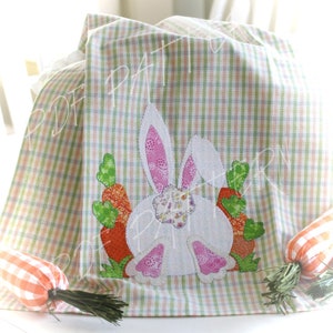 May include: A white bunny with pink ears and a floral pattern is surrounded by green and orange carrots on a green and white checkered kitchen towel.