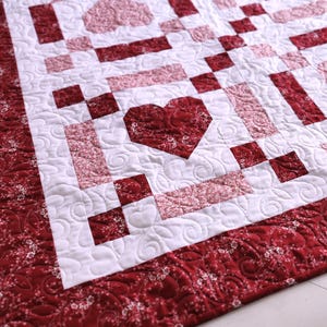 May include: A handmade quilt featuring a heart design. The quilt showcases a patchwork pattern with red and white squares and rectangles. The heart is in the center, surrounded by a floral print border. The quilt is machine quilted with a swirl pattern.