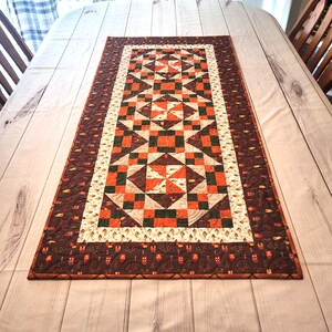 May include: A rectangular quilted table runner featuring a patchwork design of orange, dark green, and cream fabric squares. The center displays repeating star and pinwheel patterns, while the wide outer border is dark brown with small house motifs.