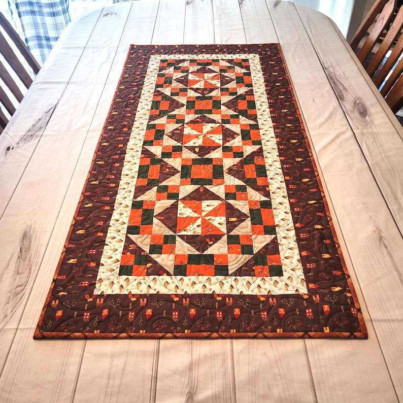 May include: A rectangular quilted table runner featuring a patchwork design of orange, dark green, and cream fabric squares. The center displays repeating star and pinwheel patterns, while the wide outer border is dark brown with small house motifs.
