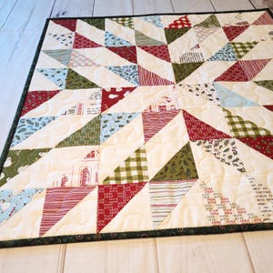 May include: A square quilt featuring a star pattern composed of triangular fabric patches in red, green, and cream. The fabric includes various holiday-themed prints, including snowmen, pine branches, and text, with a dark green border.