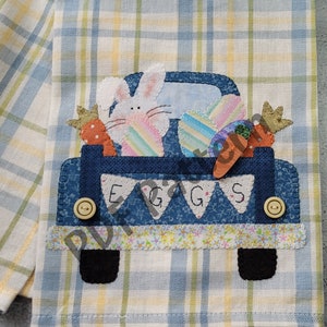 May include: A blue pickup truck with a white bunny rabbit, carrots, and colorful eggs in the bed. The truck has the word "EGGS" written on the back. The truck is on a yellow and blue plaid fabric.