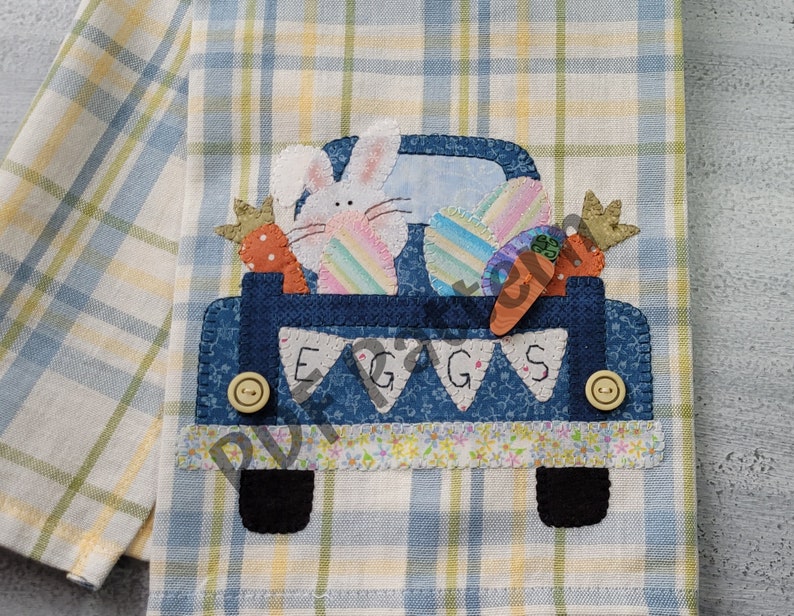 May include: A blue pickup truck with a white bunny rabbit, carrots, and colorful eggs in the bed. The truck has the word "EGGS" written on the back. The truck is on a yellow and blue plaid fabric.