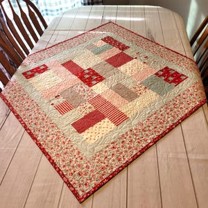 Peut inclure: Une courtepointe carr&eacute;e faite &agrave; la main avec un motif patchwork. La courtepointe pr&eacute;sente une vari&eacute;t&eacute; de tissus rouges, roses et cr&egrave;me dans un motif g&eacute;om&eacute;trique. La bordure est un imprim&eacute; floral rouge et cr&egrave;me. La courtepointe est expos&eacute;e sur une table.