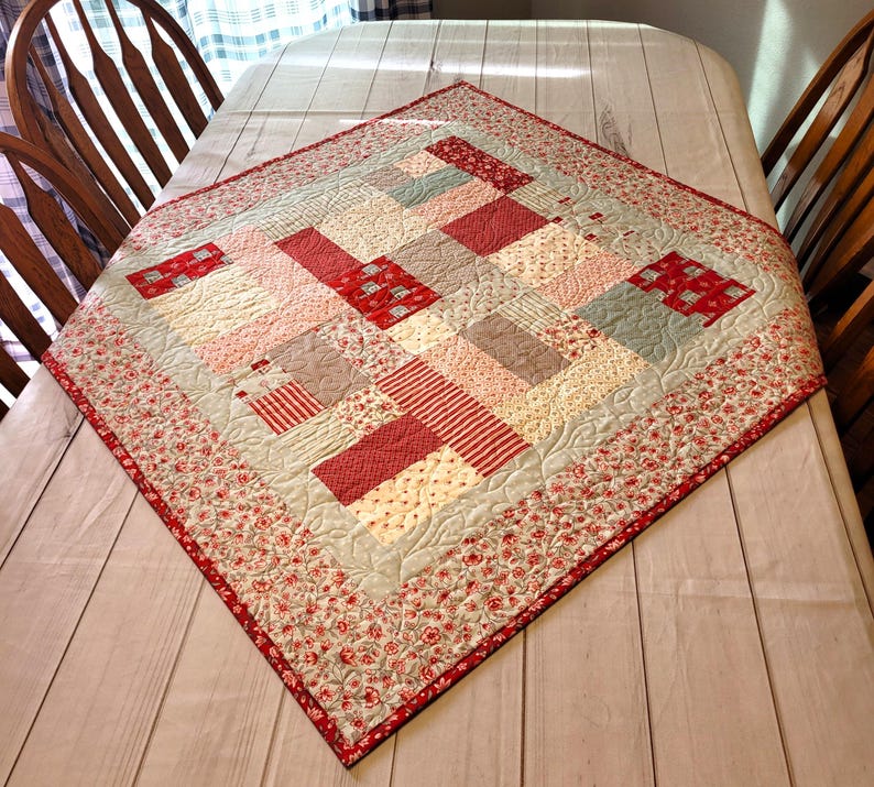 Peut inclure: Une courtepointe carr&eacute;e faite &agrave; la main avec un motif patchwork. La courtepointe pr&eacute;sente une vari&eacute;t&eacute; de tissus rouges, roses et cr&egrave;me dans un motif g&eacute;om&eacute;trique. La bordure est un imprim&eacute; floral rouge et cr&egrave;me. La courtepointe est expos&eacute;e sur une table.