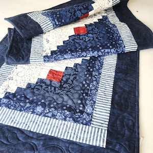 May include: A quilted table runner and placemat set in shades of blue, white, and red. The design features a log cabin pattern with floral and striped accents. The set is made of fabric and has a quilted texture.