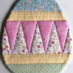 May include: A handmade quilted egg-shaped ornament with a floral pattern. The ornament is made of different fabrics in shades of blue, pink, yellow, and green.