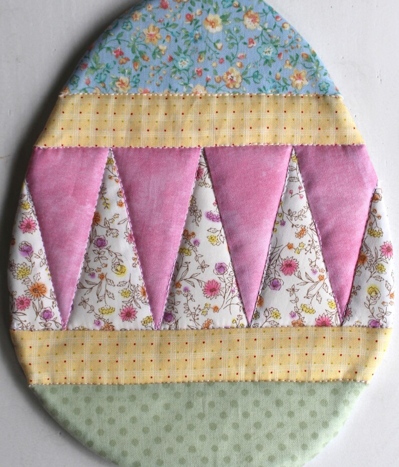 May include: A handmade quilted egg-shaped ornament with a floral pattern. The ornament is made of different fabrics in shades of blue, pink, yellow, and green.