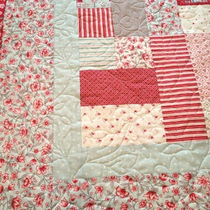 Peut inclure: Une courtepointe patchwork avec une vari&eacute;t&eacute; de motifs floraux et ray&eacute;s en rouge, blanc et bleu clair. La courtepointe pr&eacute;sente des coutures complexes et un m&eacute;lange de textures, cr&eacute;ant une sensation chaleureuse et artisanale. La courtepointe est un m&eacute;lange de motifs floraux et ray&eacute;s.