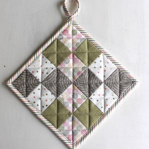 May include: A quilted pot holder with a diamond pattern in shades of green, brown, and white. The pot holder has a loop for hanging and a striped border.