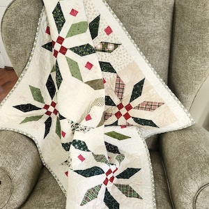 May include: A quilted blanket featuring a repeating star pattern in shades of dark green, olive, and red on a cream background. The fabric displays various small-scale prints including plaids and stars, finished with a thin green and white checkered border.