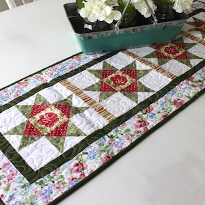 May include: A green, red, and white quilted table runner with a floral pattern. The runner has a green border and is decorated with a star pattern.